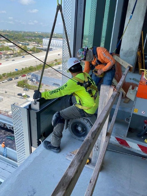 AK Glazing team members guide glazing unit suspended on cable into place at the edge of high-rise under construction 